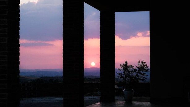 Una vista al atardecer a través de un vano con columnas, una planta en maceta y un paisaje lejano.
