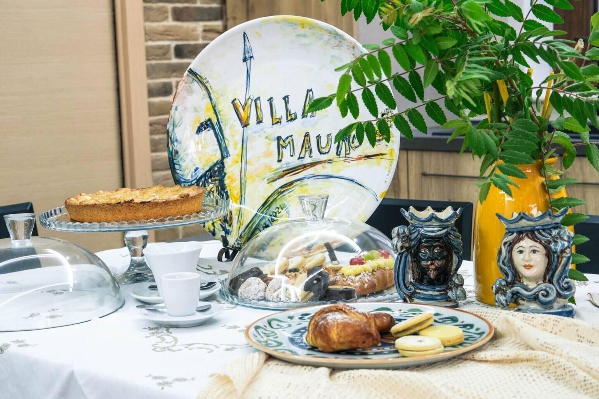Una mesa dispuesta con diversos pasteles y un pastel, un plato decorativo con el texto 'VILLA MAURO' y un jarrón amarillo con diseño de rostro.
