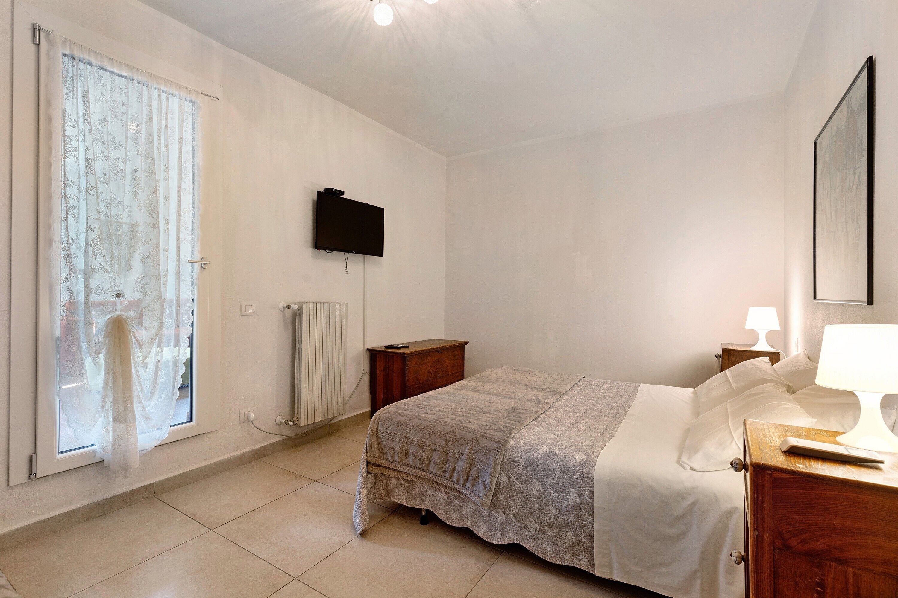 Une chambre à coucher comprenant un lit, une table de chevet, une lampe, un téléviseur fixé au mur et une fenêtre avec des rideaux.
