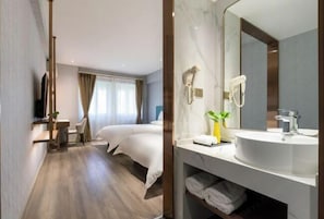 A modern hotel room with a wooden floor, a bed with white linens, a bedside table with a plant, and a large mirror above a white sink.