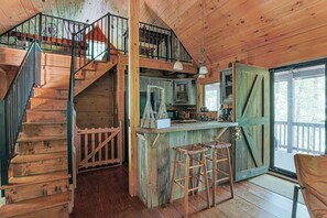 Interior rústico de una cabaña con escalera de madera, zona de cocina con barra y un gran ventanal corredero.