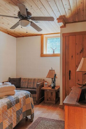 Ein gemütliches Zimmer mit Deckenventilator aus Holz, einem Fenster mit Blick, einem Holzschrank, einem kleinen Tisch mit Lampe, einer Couch und einem Bett mit Steppdecke.