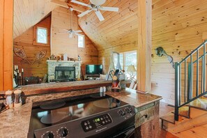 Interior acogedor de una cabaña con chimenea de piedra, una cocina con estufa, televisor y ventilador de techo.