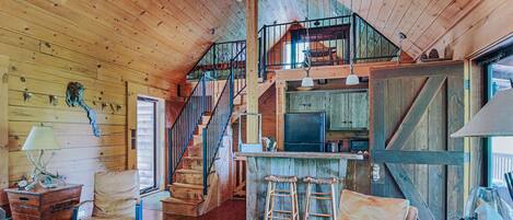 Interior rústico de una cabaña con detalles de madera, una escalera que sube a una buhardilla, una zona de cocina y una barra con taburetes.