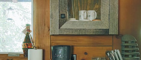 Interior de una cabaña de madera con una zona de cocina que incluye una cafetera, tazas y un aparador de vidrio con copas de vino y un pequeño adorno.