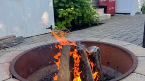 A fire pit with burning logs, surrounded by stone steps, in a backyard with a white fence and a Coca-Cola cooler in the background. - ONE Minute walk to BEACH! High end home special! TOP RATED GUEST FAVORITE (Dennis)