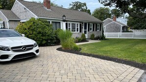 A driveway leading to a house with a white picket fence, a parked white car, and a well-maintained lawn. - ONE Minute walk to BEACH! High end home special! TOP RATED GUEST FAVORITE (Dennis)