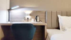 Habitación de hotel con escritorio, una silla, cama y mesita de noche con lámpara y hervidor.