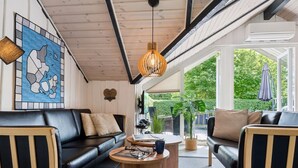 En moderne stue med en sort lædersofa, et træbord og et stort vindue med udsigt til grønne omgivelser.