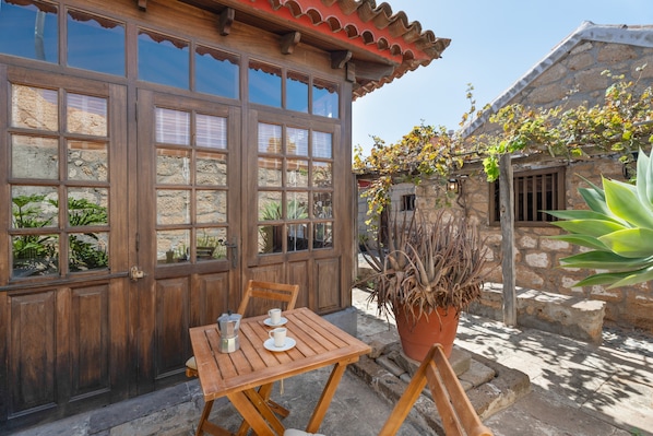 Une table en bois, avec des chaises, et un pot de fleurs, devant un bâtiment doté de grandes fenêtres et de murs en pierre.