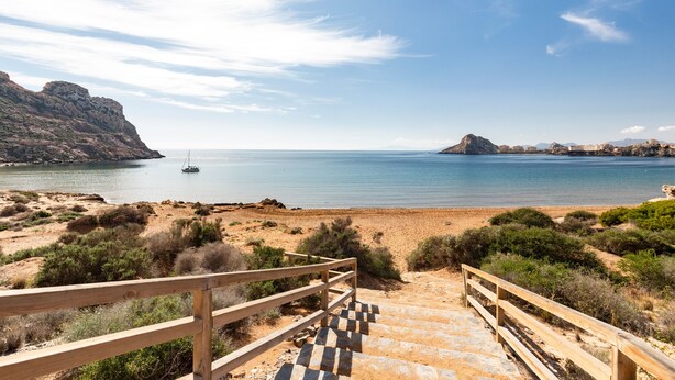 A coastal landscape with a wooden staircase leading to a beach, a sailboat on the water, and distant rocky formations. - Apartment 'Aguilas Con Ac Y Piscina' with Shared Pool, Wi-Fi and Air Conditioning (Águilas)