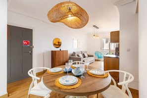 Un comedor con una mesa redonda de madera dispuesta para cuatro personas, rodeada de sillas blancas. Una lámpara colgante de fibras naturales ilumina el espacio, y un espejo está colgado en la pared.