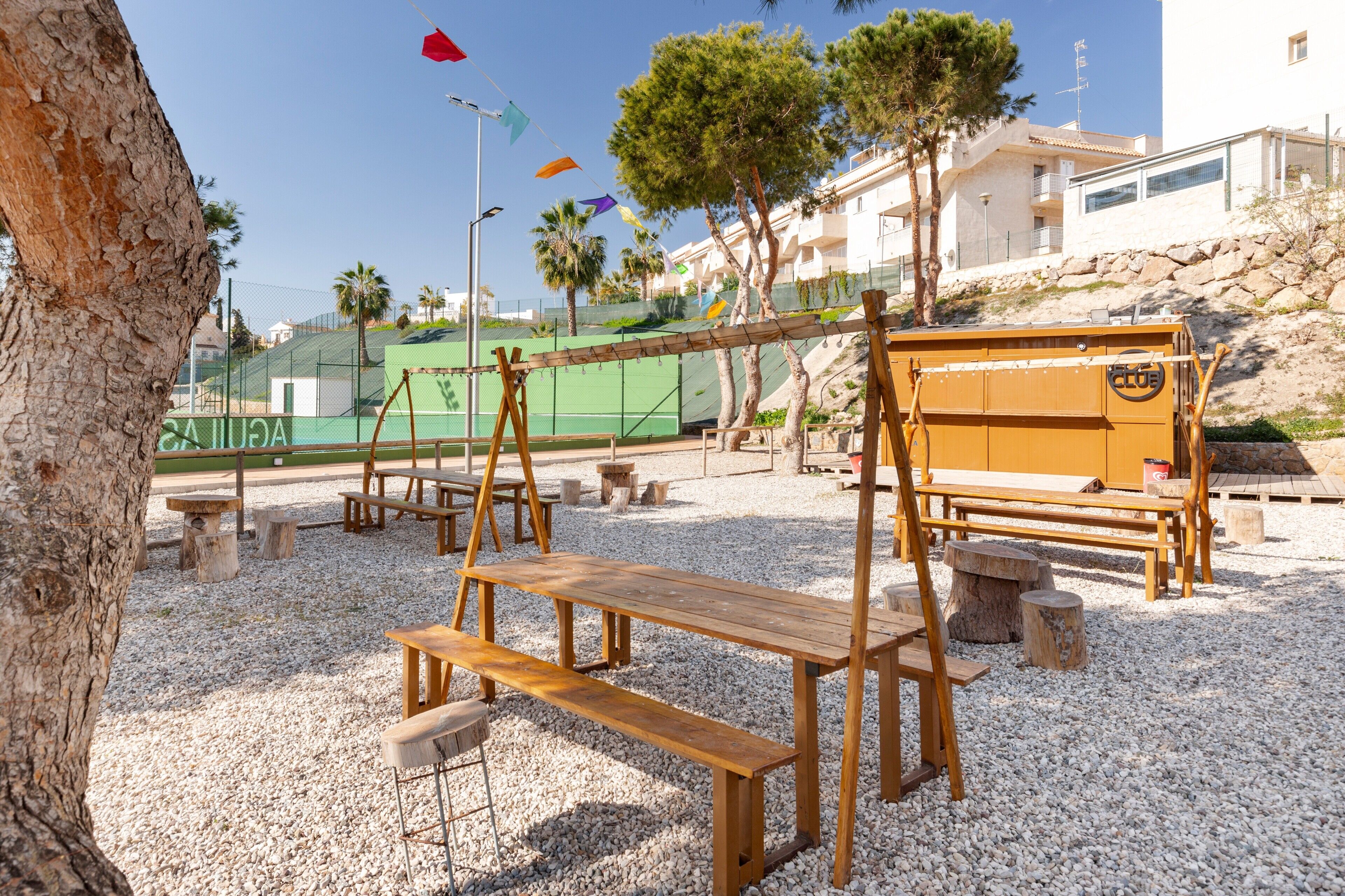 A wooden swing set, picnic tables, and benches in a gravel area with palm trees and a building in the background.