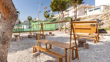 A wooden swing set, picnic tables, and benches in a gravel area with palm trees and a building in the background.