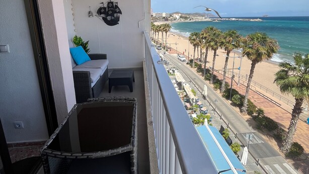 Un balcón con mobiliario de mimbre, una lámpara de techo y vista a una playa con palmeras y una isla distante.