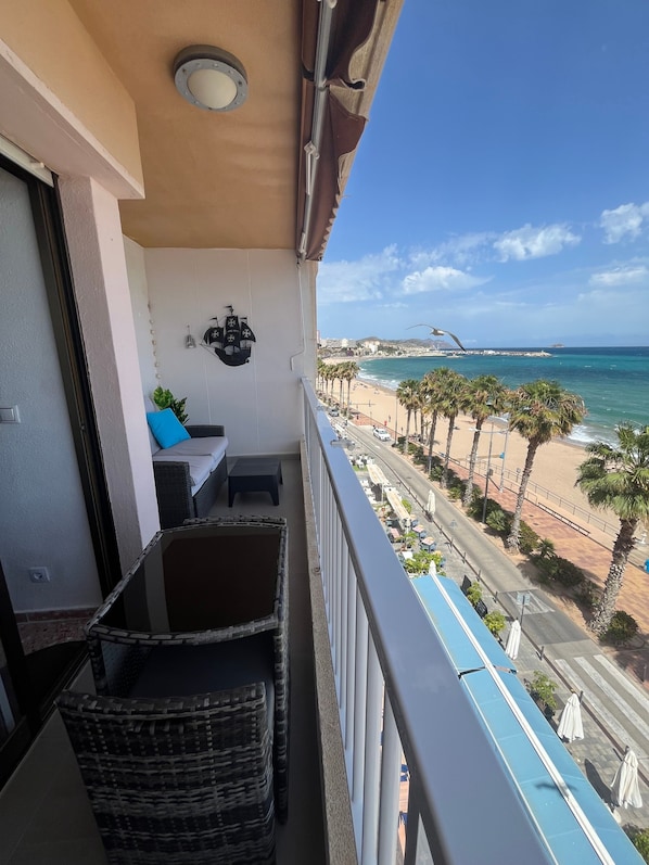 Un balcon dotĂ© de meubles en osier, dâun luminaire au plafond et offrant une vue sur une plage bordĂ©e de palmiers et dâune Ăźle lointaine.