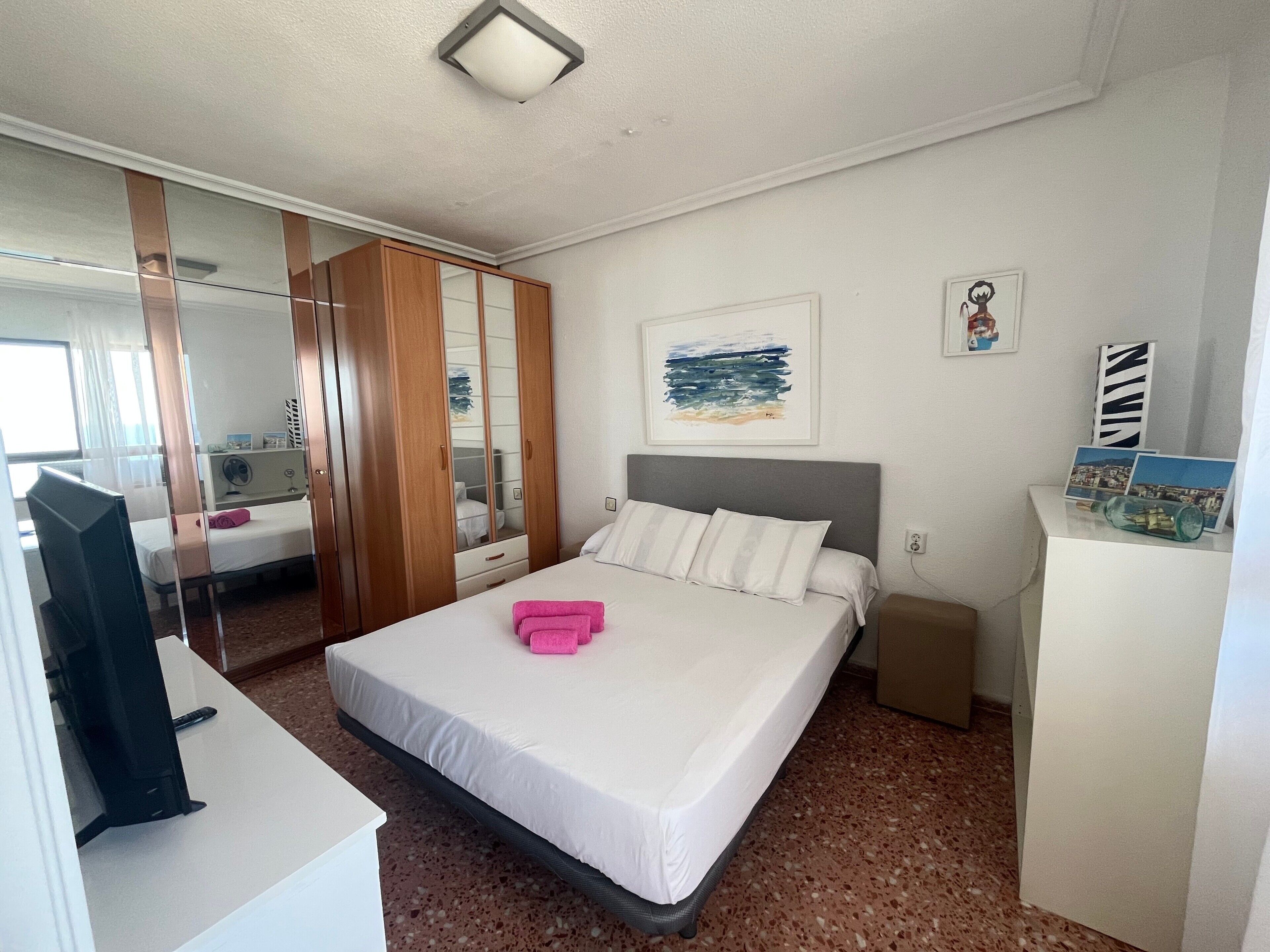 A hotel room with a bed, bedside table, a mirror, a wall-mounted artwork, and a small shelf with items.