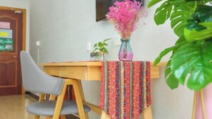 Una mesa de madera con mantel colorido, una silla gris, un florero con flores rosadas y una planta verde.