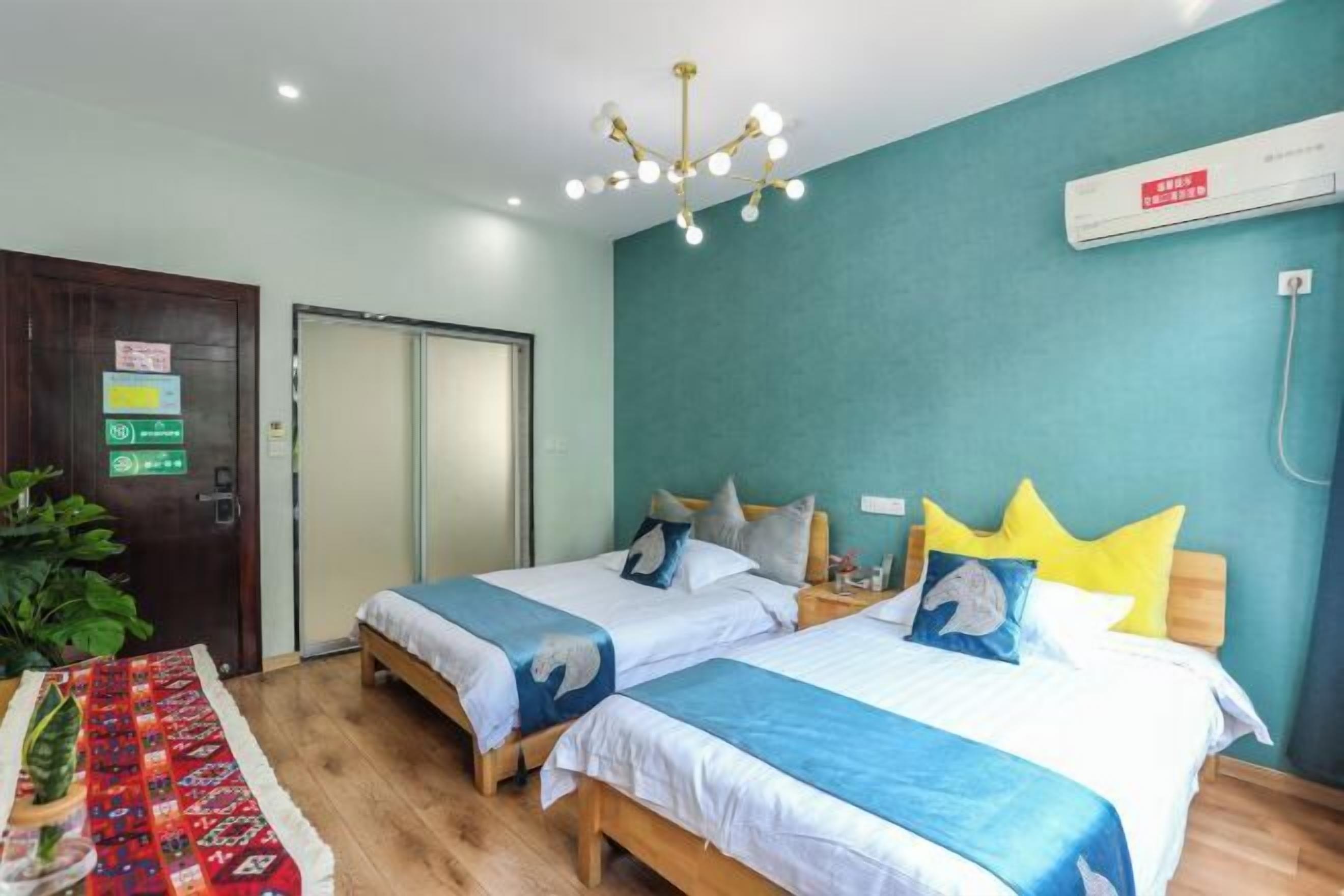 Una habitación de hotel con dos camas, suelo de madera, pared color verde azulado y una lámpara en el techo.