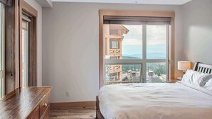 Een slaapkamer met een groot raam, een houten dressoir en een bed met witte beddengoed.