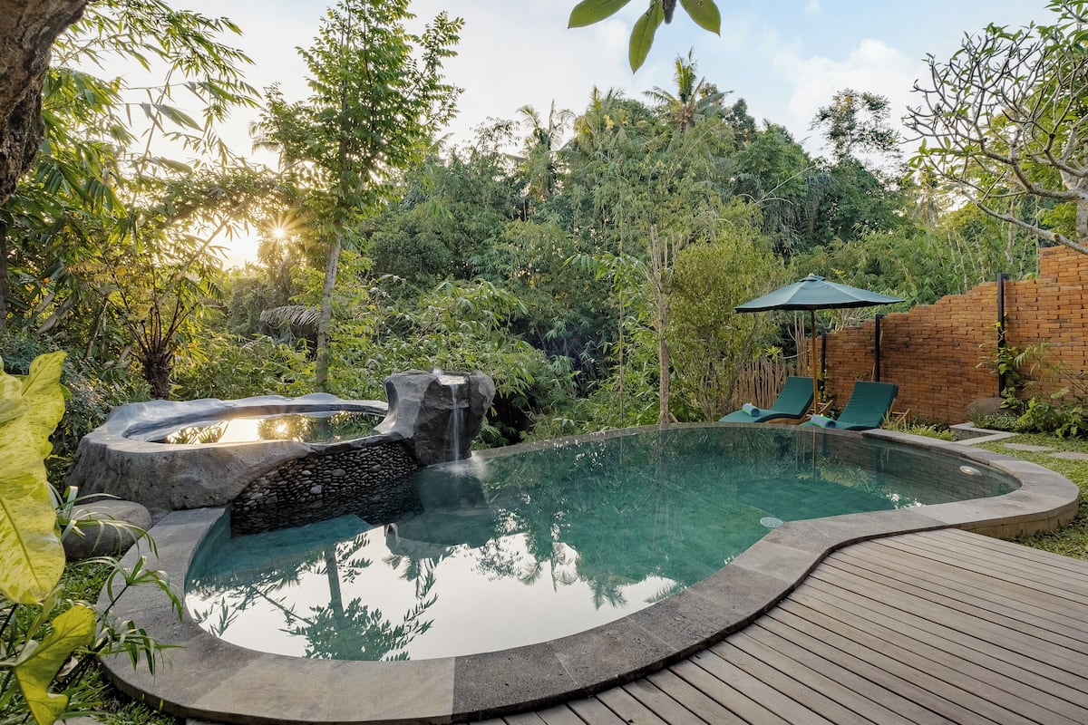 A circular hot tub with a waterfall feature, surrounded by lush greenery and a wooden deck.