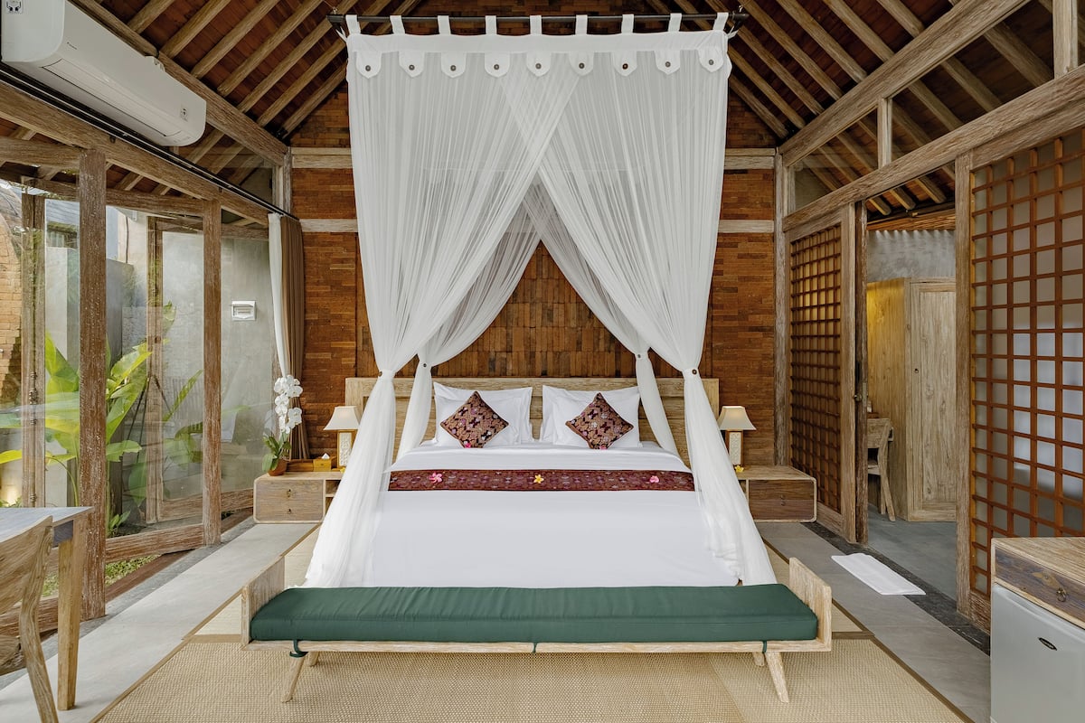 A room with a four-poster bed, wooden flooring, a green bench, a small table, and a nightstand with a lamp.