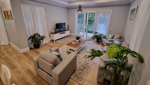 A modern living room with a sofa, coffee table, and TV stand. There are potted plants and a large window with blinds. - 229 - The Pool House (South Albury)
