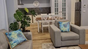 A modern living room with a grey sofa, a dining table with chairs, a large potted plant, and a kitchen area with a refrigerator and cabinets. - 229 - The Pool House (South Albury)