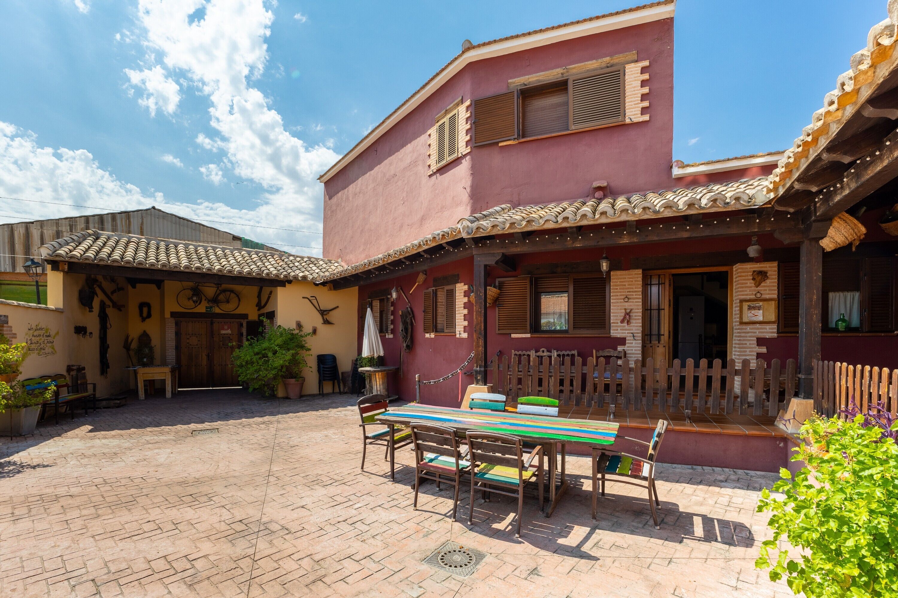 A courtyard with a tiled floor, a wooden bench, a table with chairs, and a building with a tiled roof and wooden shutters.