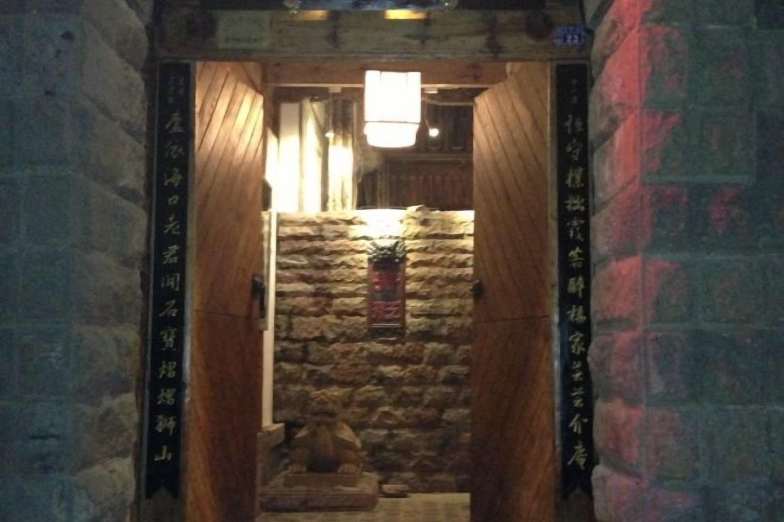 A narrow corridor with stone walls and wooden doors leading to a room with a stone wall and a hanging lantern.