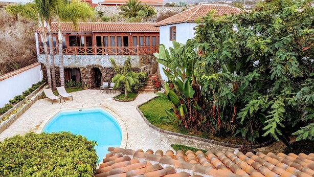 Una piscina con plataforma de piedra, un edificio blanco con techo rojo y abundante vegetación.