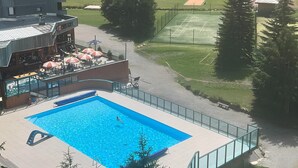 A swimming pool with a person swimming, a covered outdoor seating area, and a sports field in the background.