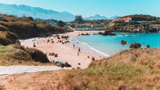 Una playa costera con cielo despejado, costa rocosa y montañas a lo lejos.