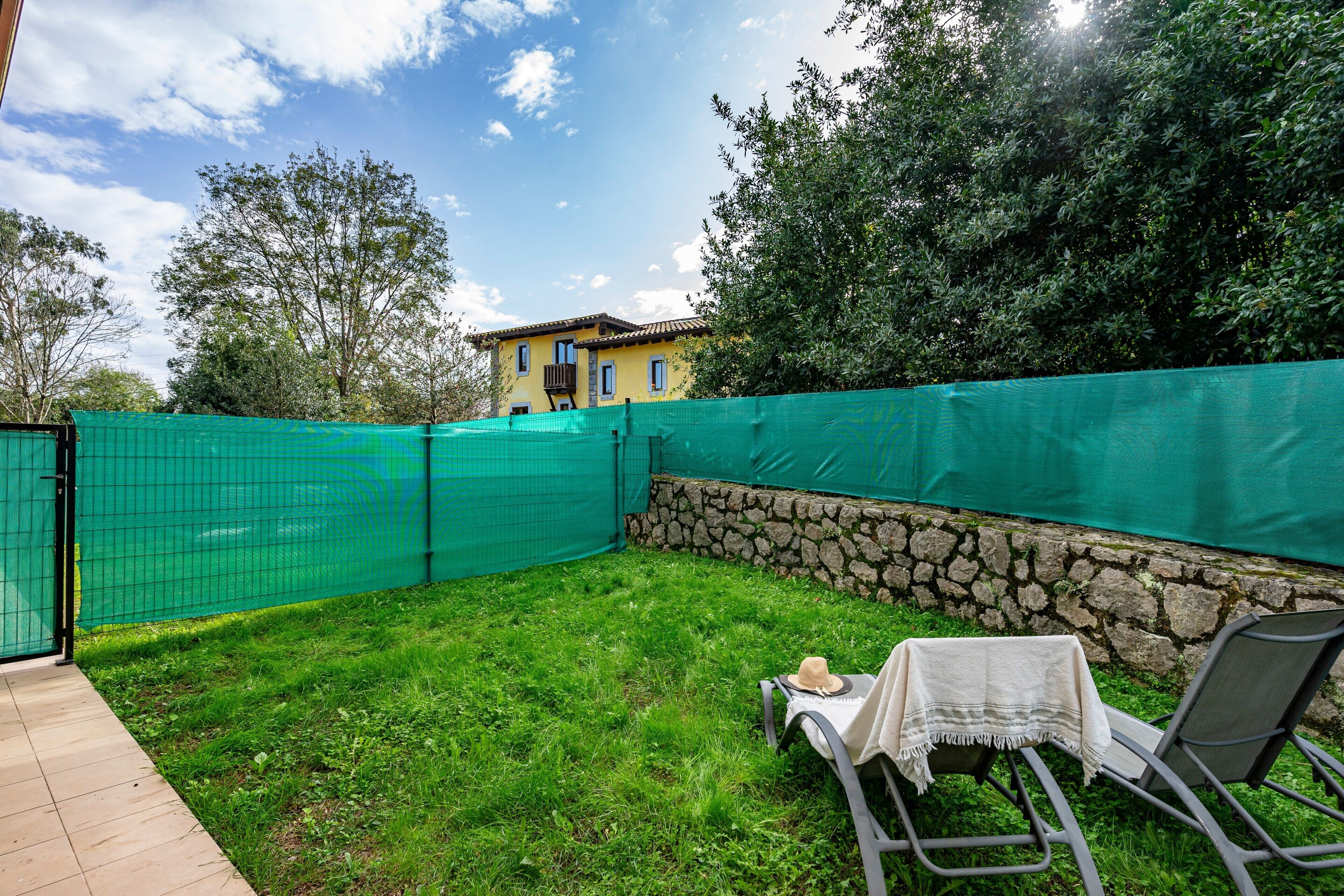 A backyard with a green fence, a stone wall, and a house in the background.
