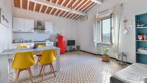 Een kamer met een eettafel, stoelen, een keukenhoek, een televisie en een raam met uitzicht.