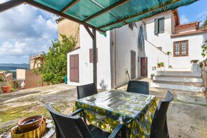 Une terrasse couverte avec une table et des chaises, un escalier et un bâtiment blanc avec des volets.