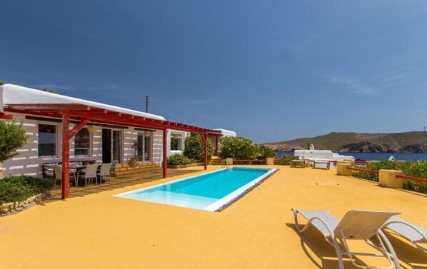 Zona de piscina con una pérgola roja, sillones blancos y vista al mar.