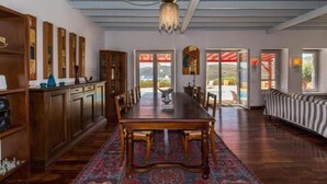 A spacious room with a long wooden table, a chandelier, and a view through large windows. - Villa 'Agios Sostis Mykonos' with Sea View, Wi-Fi and Air Conditioning (Mýkonos City)