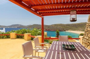 Una terraza con sillas de mimbre y una mesa, con vista a un paisaje costero que ofrece una vista despejada del mar y colinas distantes.