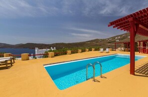 Una piscina con un pérgola roja, sillas de descanso y vistas al mar y colinas.