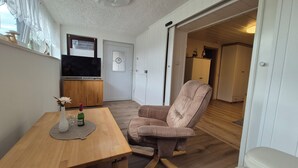 Uma sala de estar com uma mesa de madeira, uma poltrona marrom, uma televisão sobre um armário de madeira e uma porta que dá para outro cômodo.