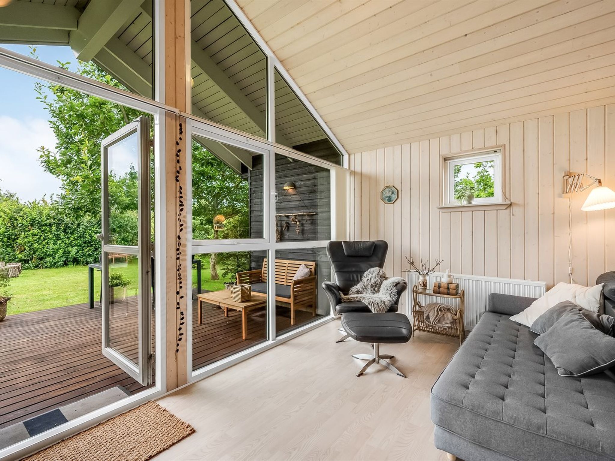 En moderne stue med et stort glasvindue der fører ud til en terrasse, en grå sofa, en sort lænestol og et lille bord med en lampe.