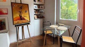 Ett rum med ett glasbord, två stolar, ett staffli med en målning, hyllor med diverse föremål och ett fönster med utsikt över träd.