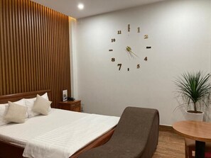 Une chambre d’hôtel avec un lit, une table de chevet, une horloge murale, une petite table et une plante en pot.