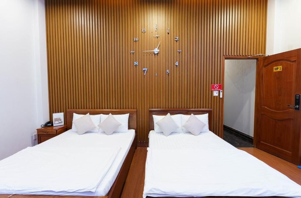 Una camera d'albergo con due letti, una sveglia a muro in legno, una porta con la scritta 'VIP 1' e un tavolino con un telefono e una cornice.