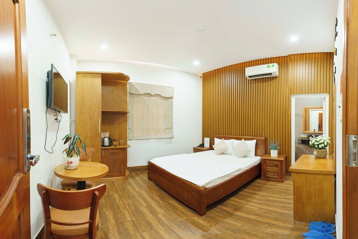 Habitación de hotel con una cama de madera, un escritorio con una silla, una mesita con una planta y un aire acondicionado instalado en la pared.
