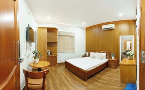 Ein Hotelzimmer mit einem hölzernen Bett, einem Schreibtisch mit Stuhl, einem kleinen Tisch mit Pflanze und einer Wand-Klimaanlage.
