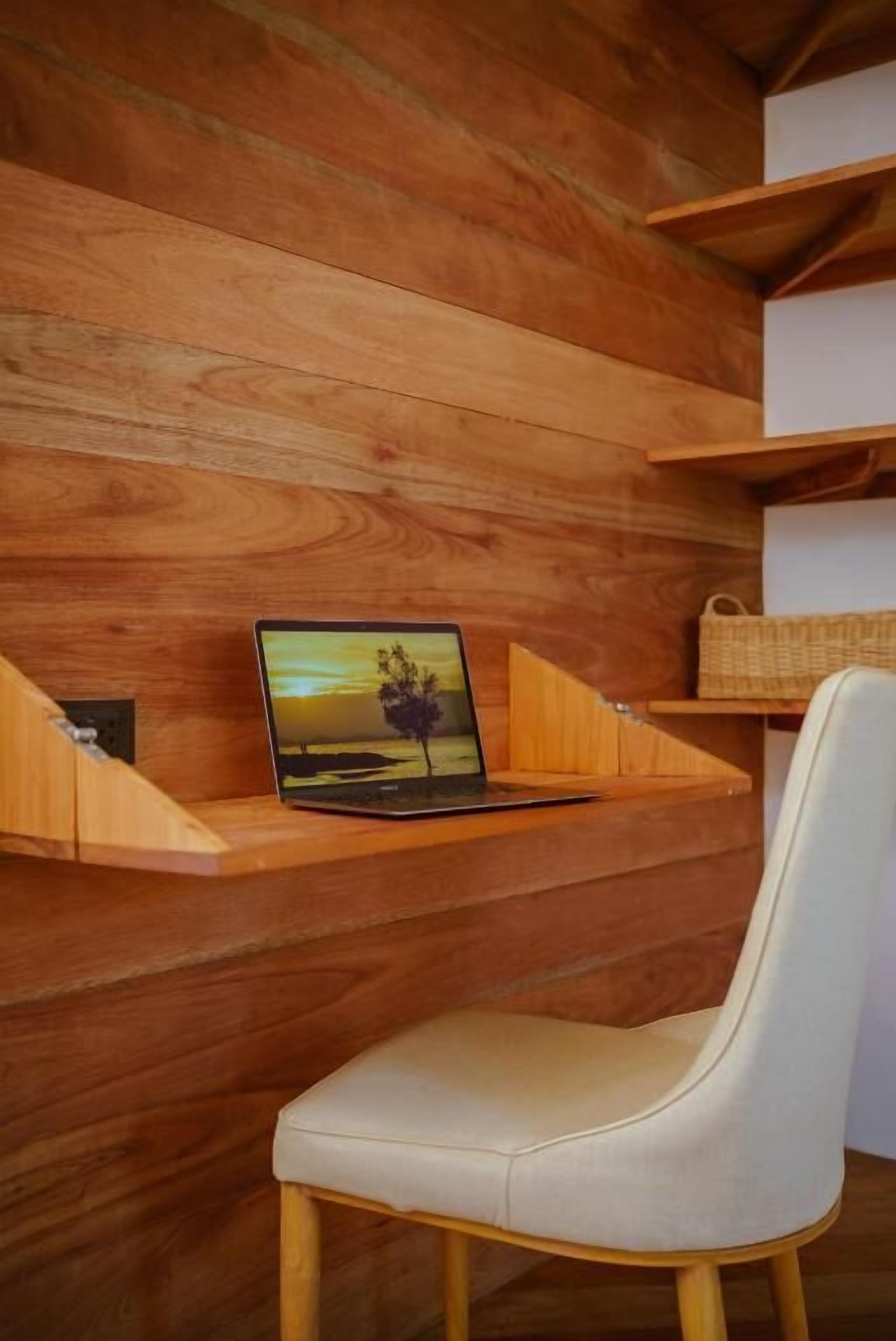Un escritorio de madera con una laptop, una silla blanca y una cesta tejida sobre estantes.