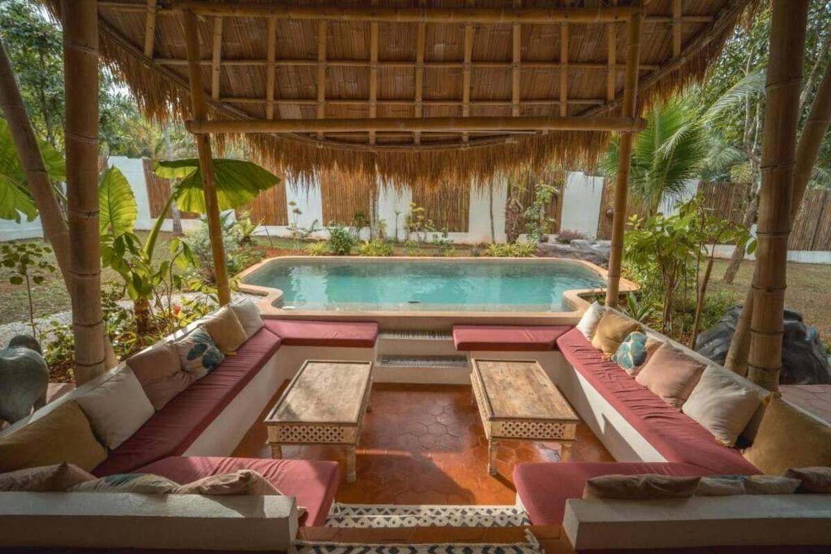 A pool area with a thatched roof, seating, and tropical plants.