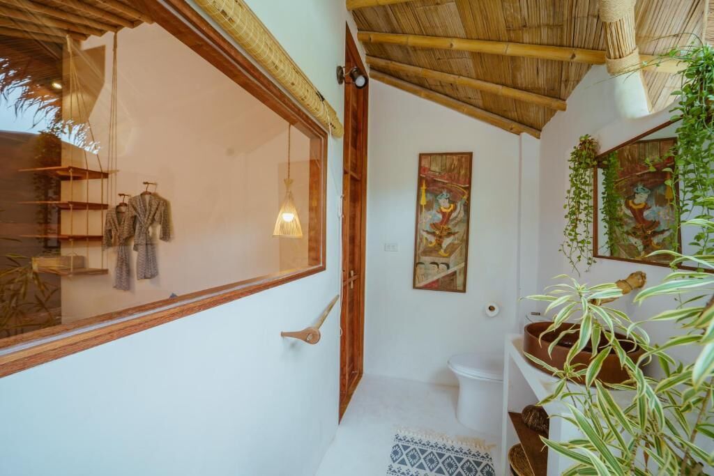 Un baño con puerta de madera, un inodoro, un estante con toallas y una ventana que da a una habitación con ropa colgada.